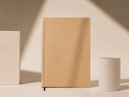 A collage of four warm-toned studio shots showing a tan hardcover notebook or journal with white geometric props. One panel shows the closed notebook standing upright, another shows it closed on stacked white blocks, a third shows it open with blank white pages, and a fourth shows a close-up of the closed cover and black ribbon bookmark.